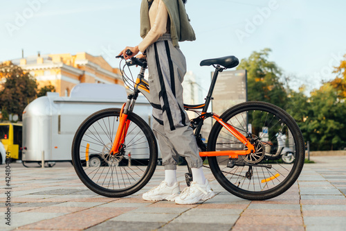 Cropped photo of a modern colorful bright orange bike and woman's legs in stylish pants and white training shoes in urban scenery. Details.
