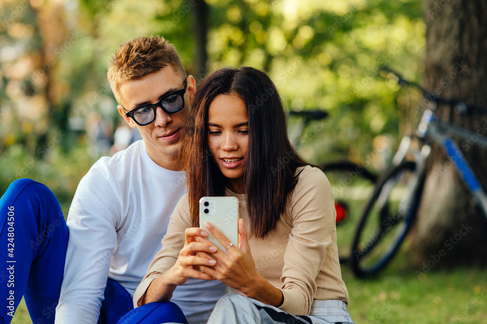 Pretty hispanic female is holding a smartohone, texting, showing screen to her boyfriend. The couple is watching on the phone, sitting in the park.