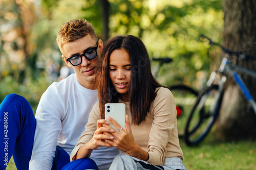 Pretty hispanic female is holding a smartohone  texting  showing screen to her boyfriend. The couple is watching on the phone  sitting in the park.