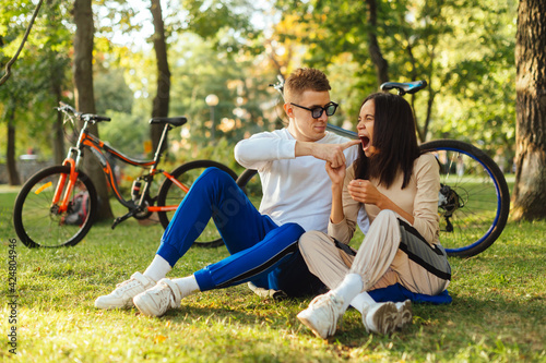 Funny young couple is laughing and joking, when sitting on a grass in the park. Two people having fun, a man is pointing a finger to a woman's mouth, a female is trying to bite a finger.