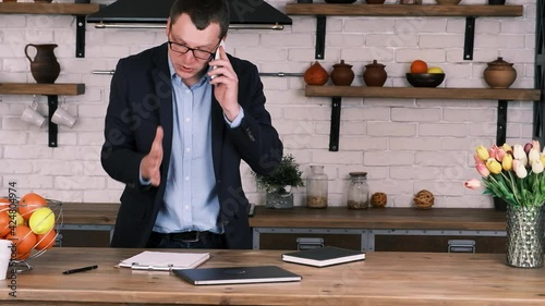Disgruntled aggressive entrepreneur in glasses and business suit is actively gesturing while talking on cell phone while standing in kitchen. He throws out phone on table and pushes notebook off table