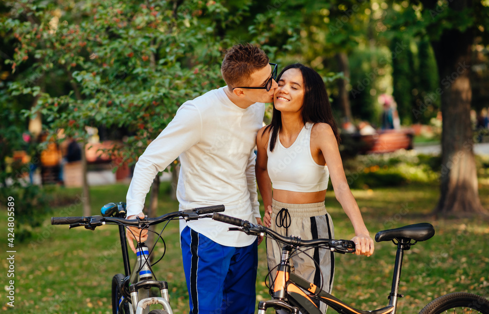Obraz premium Lovely couple standing with their bikes in the park. Attractive romantic guy is kissing his girlfriend on the cheek, the woman is smiling with her eyes closed.