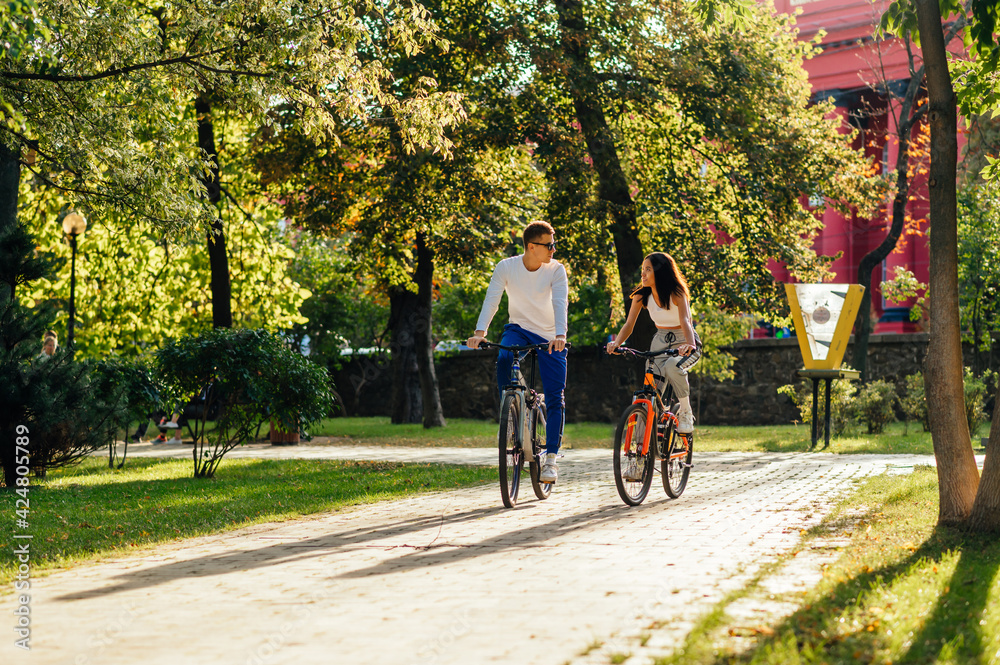Two young bikers on a modern colorful bicycles are riding outside, talking to each other, having fun on a sunny day. Couple in casual outfits is having rest.