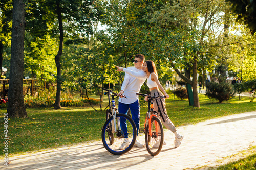 Two young adults are in the park with their bicycles. Attractive man in sunglasses is pointing his finger, showing something to the concentrated woman.
