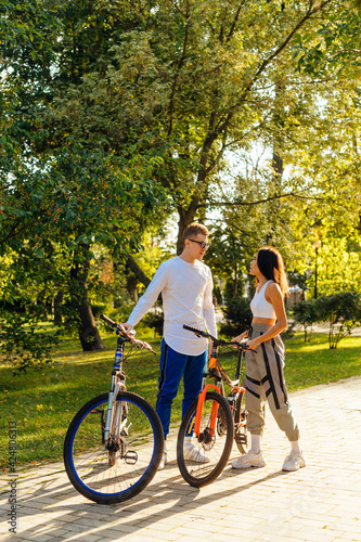 Vertical shot of a girlfriend and boyfriend having a conversation in the city park, standing with colorful bicycles. Attractive young couple is talking.