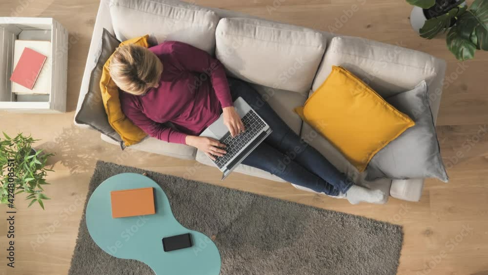 woman lying on couch typing on laptop computer top down view,,comfortable female resting on sofa wearing casual clothing using notebook writing email works from home high angle shot Vertical 9 16