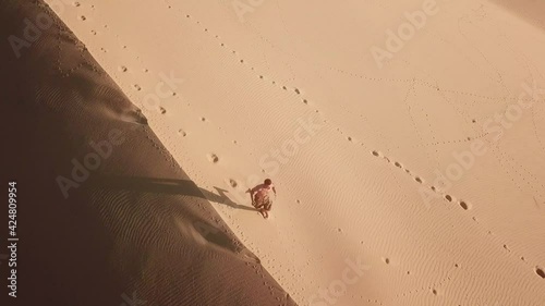 Aerial view of man running along the large sand dunes. Drone point view of a young man running in the desert at sunrise.
