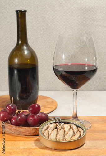 Top, front view, medium distance of a glass of red wine, bottle of wine, corkscrew, artisan bowl of red grapes, and a tin of sardines in olive oil, on rare wood plate