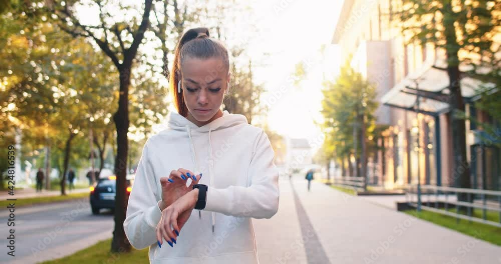 Strong female athlete using stop watch to time jogging on smart digital gadget. Young Caucasian woman setting up device and running along modern buildings in city street. Sport, health concept.