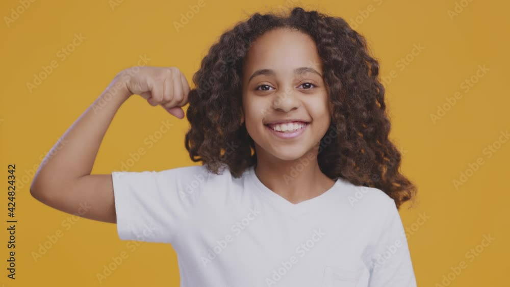 Little and strong. Positive black girl smiling and showing her biceps, orange background Little and strong. Positive black girl smiling and showing her biceps, orange background