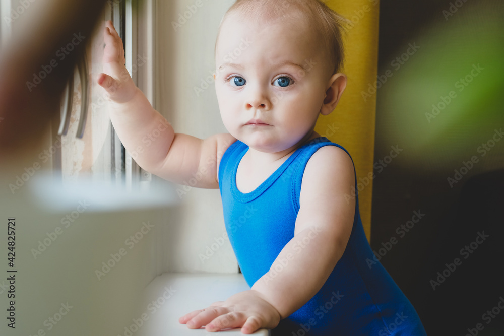 Lockdown due to COVID-19 concept. Portrait of sad pensive caucasian toddler boy standing near window at home. Lockdown due to COVID-19 concept. Portrait of sad pensive caucasian toddler boy standing near window at home.