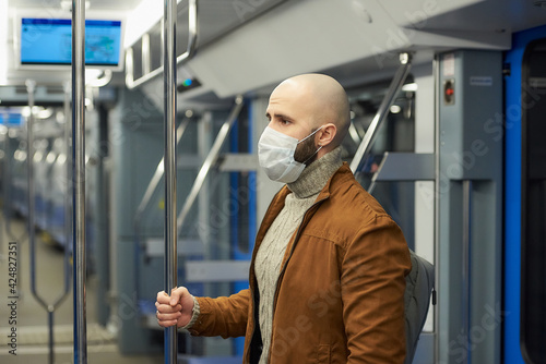 A man with a beard in a medical face mask to avoid the spread of coronavirus is standing and holding the handrail in a subway car. A bald guy in a surgical mask is keeping social distance on a train.