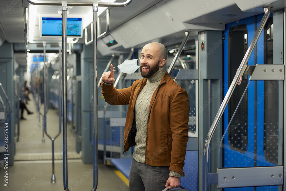 A man with a beard is taking off a medical face mask and smiling in a subway car. A bald guy with a surgical mask is keeping social distance on a train.