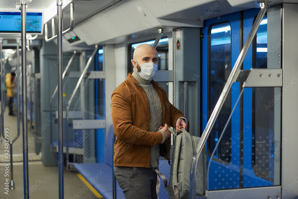 A man with a beard in a medical face mask to avoid the spread of coronavirus is putting on a backpack while riding a subway car. A bald guy in a surgical mask is keeping social distance on a train.