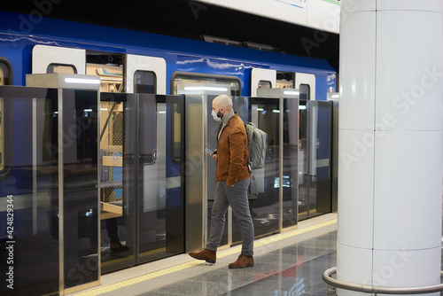 A man in a medical face mask to avoid the spread of coronavirus is holding a cellphone while entering the modern subway car. A bald guy in a surgical mask is keeping social distance.