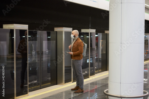 A full-length photo of a man in a face mask is holding a smartphone while waiting for a train at the subway platform. A bald guy in a surgical mask is keeping social distance.