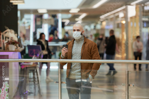 A man in a face mask to avoid the spread of coronavirus is holding a cup of coffee in the shopping center. A bald guy in a surgical mask is keeping social distance.