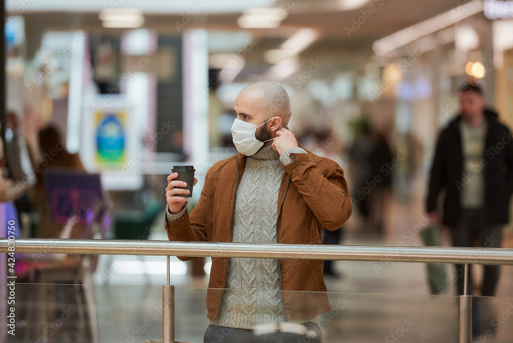 A man with a beard is putting on a medical face mask while holding a brown cup of coffee in the shopping center. A bald guy is keeping social distance.