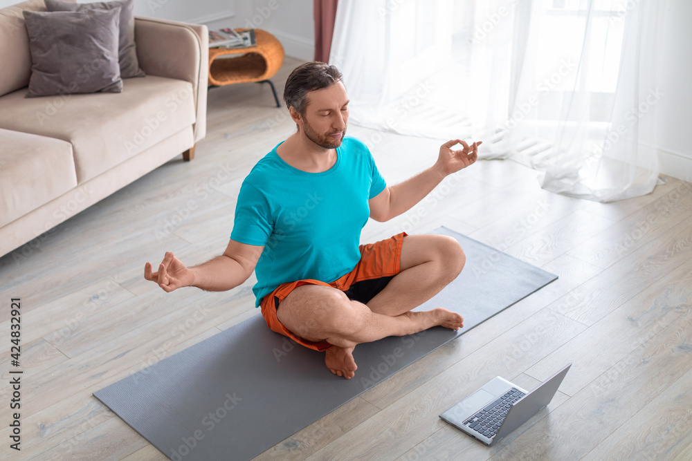 Man taking an online yoga class. He is meditating sitting on the floor in front of a laptop monitor.