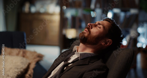 Tired young businessman returning home removing suit tie relaxing on the sofa. Living room apartment. Corporate person ;lifestyle. Workaholic.