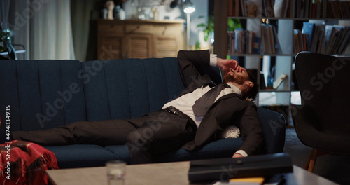 Very hardworking young businessman arriving home after difficult work resting on sofa lying down. Workaholic people. Overworking is bad for health.