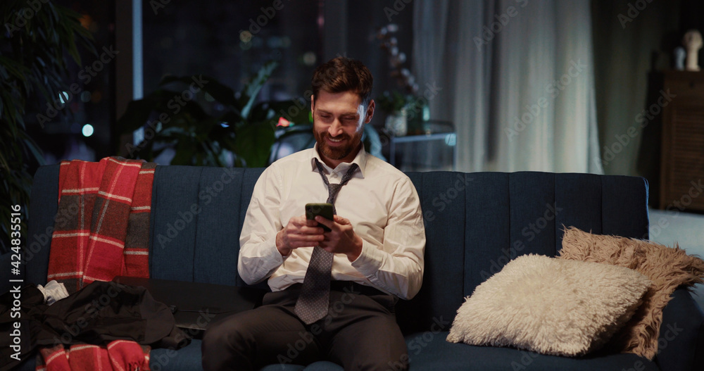 Obraz premium Happy adult young business person in suit sitting on sofa relaxing at home using application social network on smartphone screen networking online.