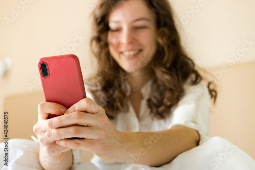 Close up modern smartphone in red case in woman hands, smiling female user sitting in bed at home. Ordering food delivery, making video call, browsing website, checking email,chatting, booking, taxi 