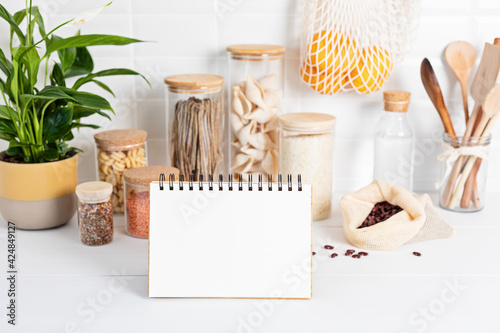 Blank notebook and assortment of grains  cereals and pasta in glass jars and kitchen utensils on wooden table. Sustainable lifestyle  zero waste storage  eco friendly idea