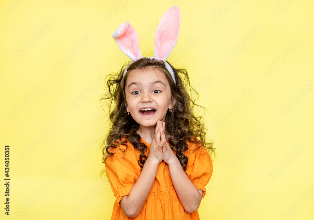 Happy curly little girl wears bunny ears, loves the celebration and mystery of the holiday, dressed in orange bright dress, isolated on yellow background. Seasonal holiday concept 