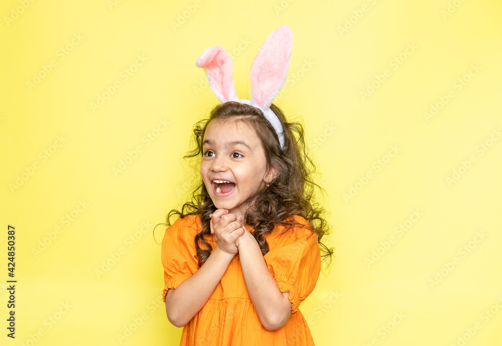 Happy curly little girl wears bunny ears, loves the celebration and mystery of the holiday, dressed in orange bright dress, isolated on yellow background. Seasonal holiday concept 