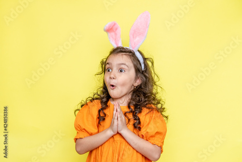 Happy curly little girl wears bunny ears, loves the celebration and mystery of the holiday, dressed in orange bright dress, isolated on yellow background. Seasonal holiday concept 