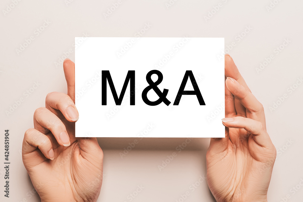 Female hands hold card paper with text M and A on a yellow background. Business and finance concept Female hands hold card paper with text M and A on a yellow background. Business and finance concept