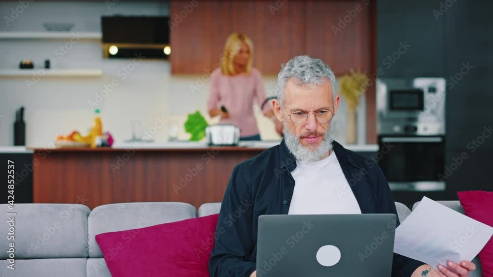 Handsome gray-bearded man working with laptop while sitting on sofa in studio kitchen. Woman preparing food standing in the background Handsome gray-bearded man working with laptop while sitting on sofa in studio kitchen. Woman preparing food standing in the background