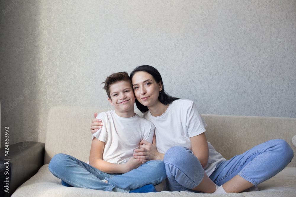 a happy brunette mom in a white T-shirt and jeans hugs her loving son sitting at home on the couch. a happy brunette mom in a white T-shirt and jeans hugs her loving son sitting at home on the couch.