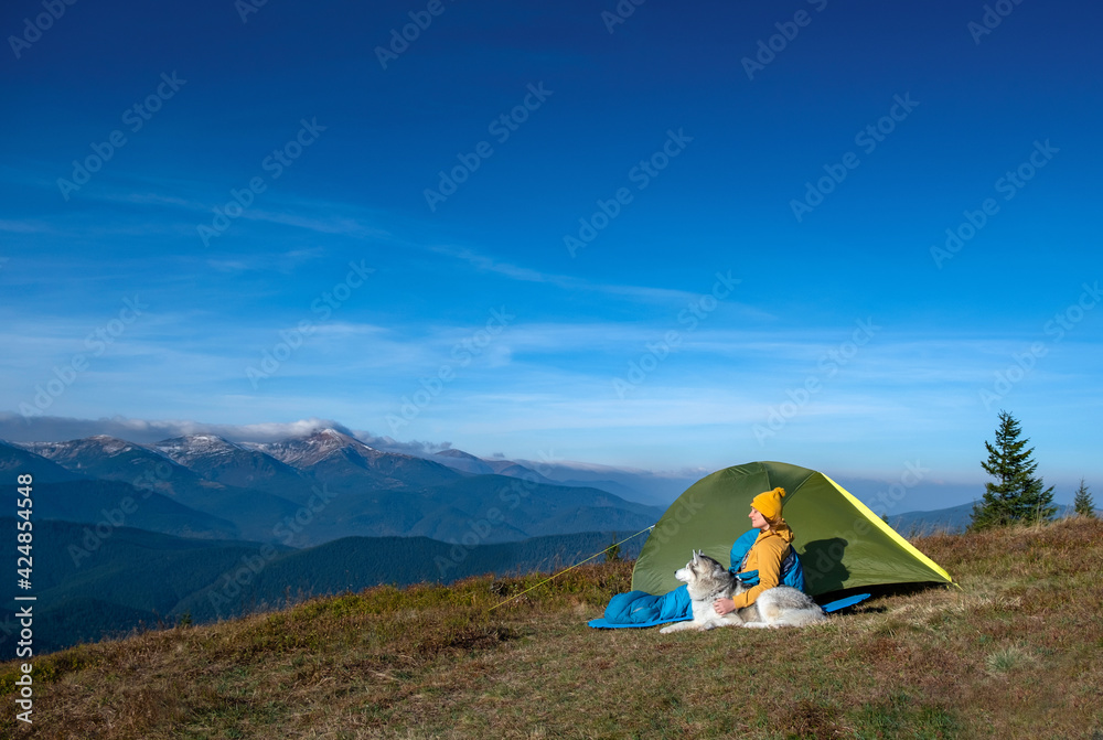 A young girl with dog is sitting with sleeping bag near the tent with mountains on background