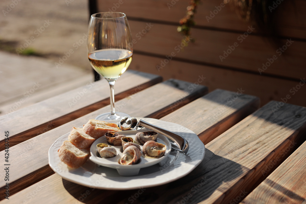 Glass with wine and dish with the snail shells, served on a white classic plate, with various sauces, pesto, truffle, cheese, pieces of French baguette, fork for a shell, wooden table in outdoor cafe Glass with wine and dish with the snail shells, served on a white classic plate, with various sauces, pesto, truffle, cheese, pieces of French baguette, fork for a shell, wooden table in outdoor cafe