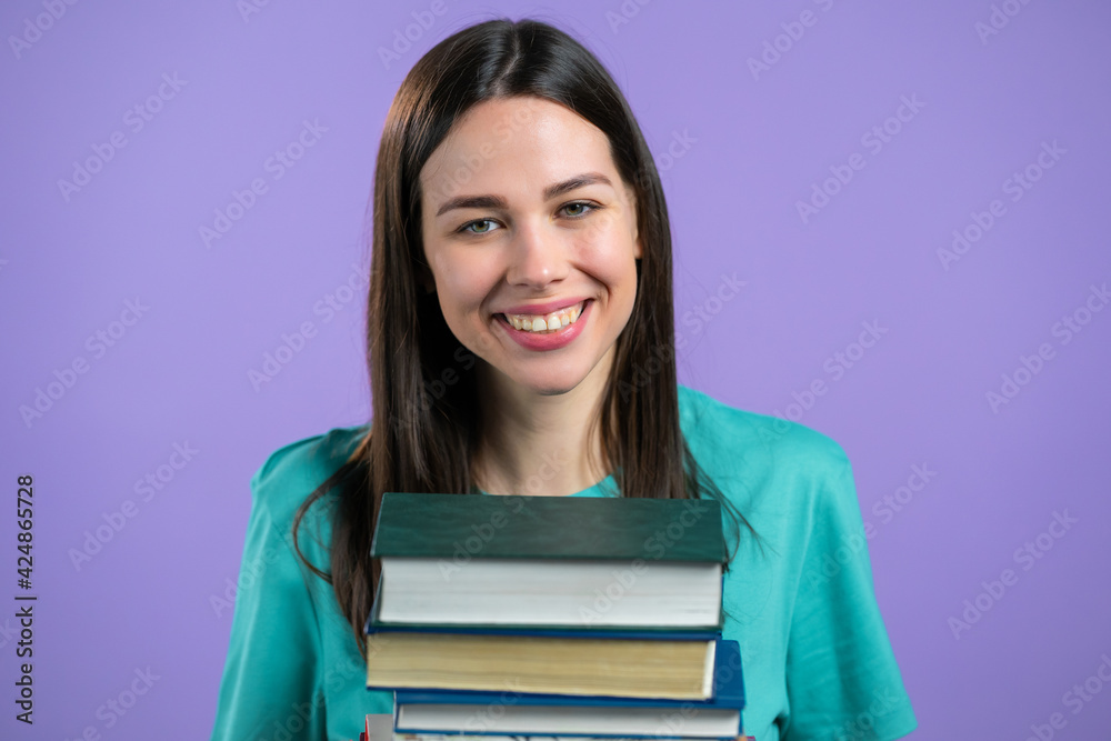 Student holds stack of university books from library on violet background in studio. Woman smiles, she is happy to graduate. Student holds stack of university books from library on violet background in studio. Woman smiles, she is happy to graduate.