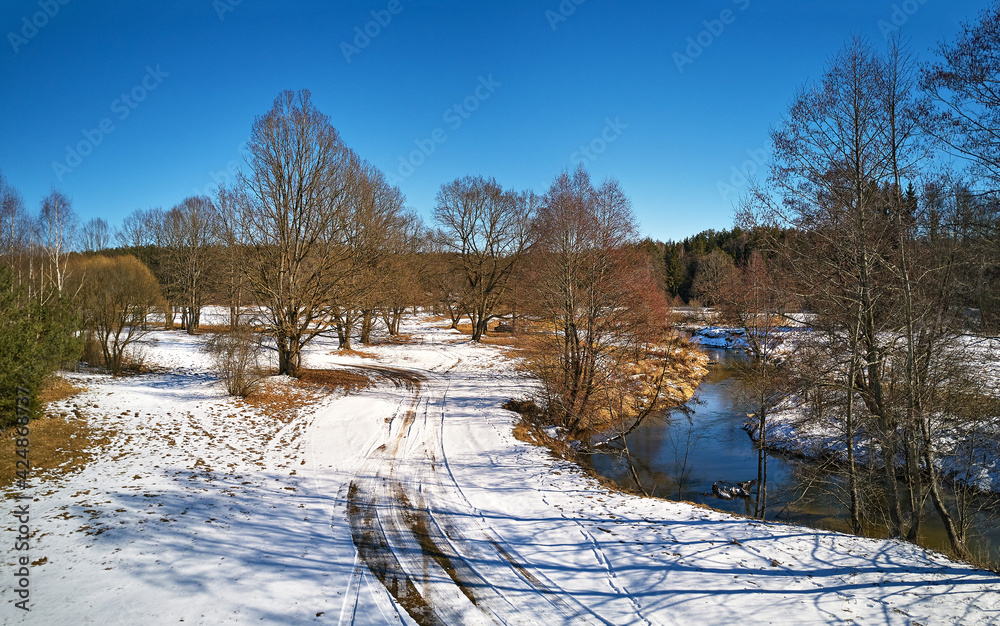 Early spring landscape with melting snow. Dirt rural road, wood, ice and snow panorama.