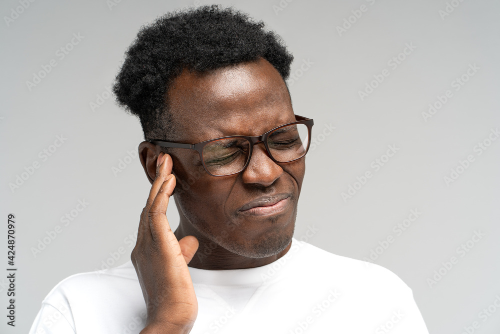 Fototapeta premium Sick African American millennial man suffering from tinnitus, throbbing earache, tired of noise. Irritated black frowning male in glasses touching painful ear, isolated on grey studio background.