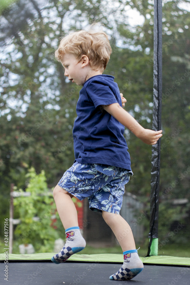 Fototapeta premium A cute little boy with blonde hair and blue eyes wearing a blue T-shirt and short shorts jumping on a trampoline. In the background, green leaves of trees and other vegetation.