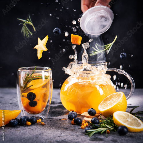 Transparent glass cup and teapot with yellow fruit tea, on a gray background. Exotic ingredients fall into the teapot with a splash.