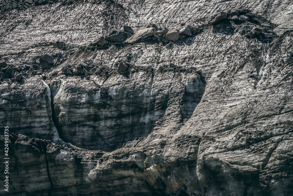 Naklejka premium Nature background with icefall near glacier with cracks and scratches. Natural backdrop with icy wall and moraines. Beautiful landscape with shiny glacial wall and stones in sunlight. Glacier texture.