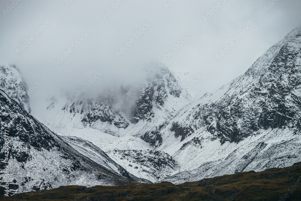 Foggy mountain landscape with white snow on black rocks in cloudy weather. Misty mountain minimalism of snowbound mountain in low clouds. Minimalist nature background of snowy mountainside in fog.
