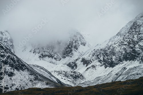 Foggy mountain landscape with white snow on black rocks in cloudy weather. Misty mountain minimalism of snowbound mountain in low clouds. Minimalist nature background of snowy mountainside in fog.