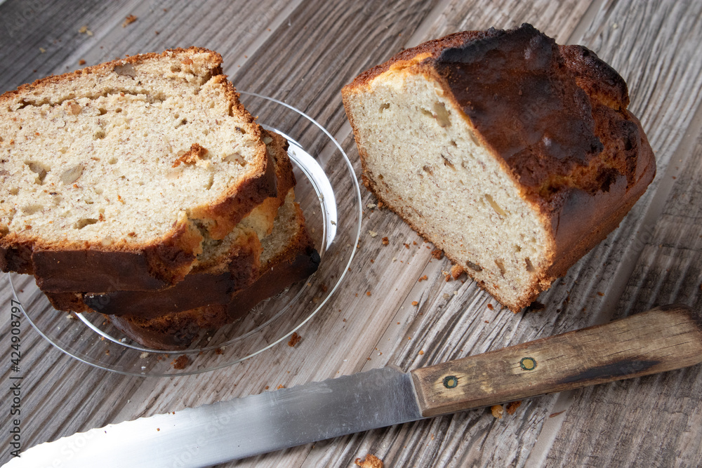 Slices of banana nut bread on a clear plate on a wood table. A loaf of bread and knife site on the table.