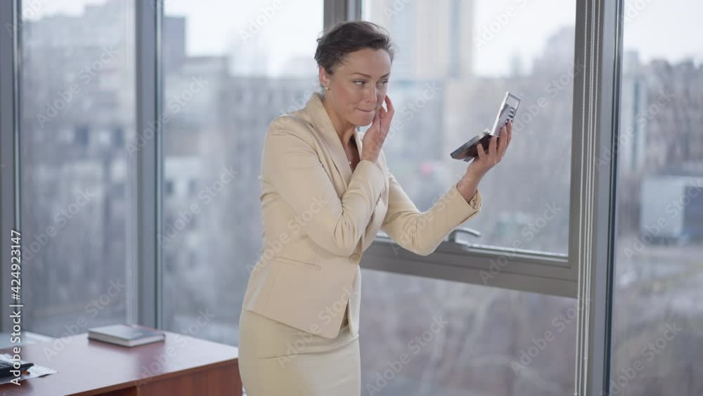 Slim gorgeous Caucasian woman adjusting make-up standing at floor-to-ceiling window in office. Confident beautiful CEO manager employee indoors at workplace. Femininity and business Slim gorgeous Caucasian woman adjusting make-up standing at floor-to-ceiling window in office. Confident beautiful CEO manager employee indoors at workplace. Femininity and business
