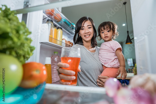asian mother carrying her daughter while taking a glass of juice inside the fridge