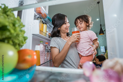 asian mother carrying her daughter while taking a glass of juice inside the fridge