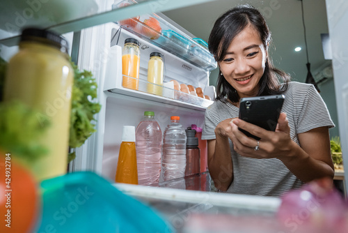 Konstfotografi asian young woman using her smartphone to buy groceries while open her fridge at