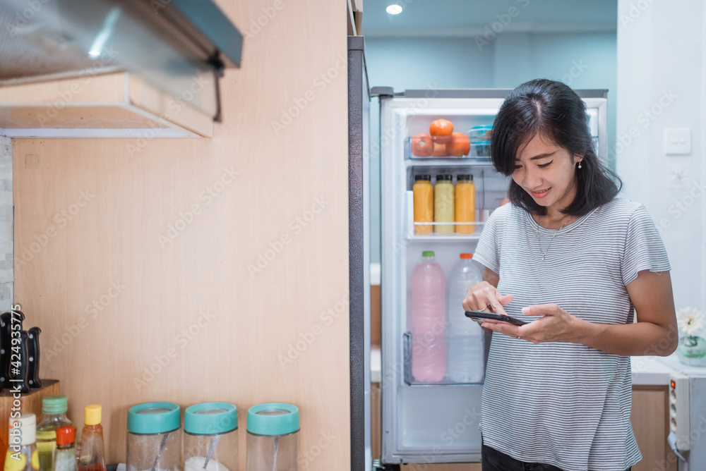 asian young woman using her smartphone to buy groceries while open her fridge at home